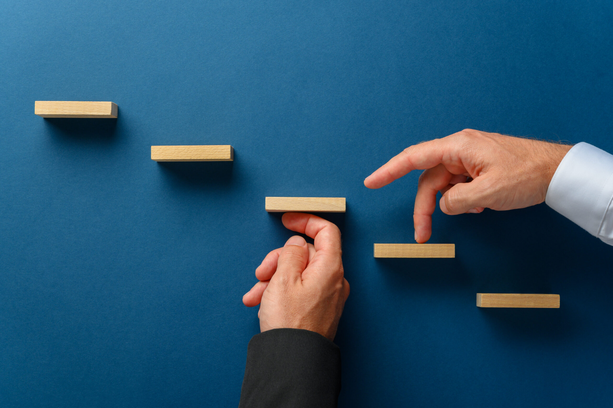 Hand of a businessman supporting wooden steps for his partner to walk his fingers upwards in a conceptual image of business partnership. Over blue background.